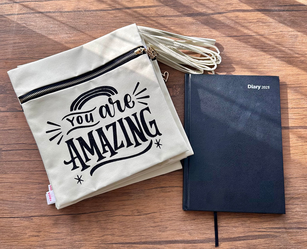 Beige pouch with 'You are Amazing' text and black notebook on a wooden surface