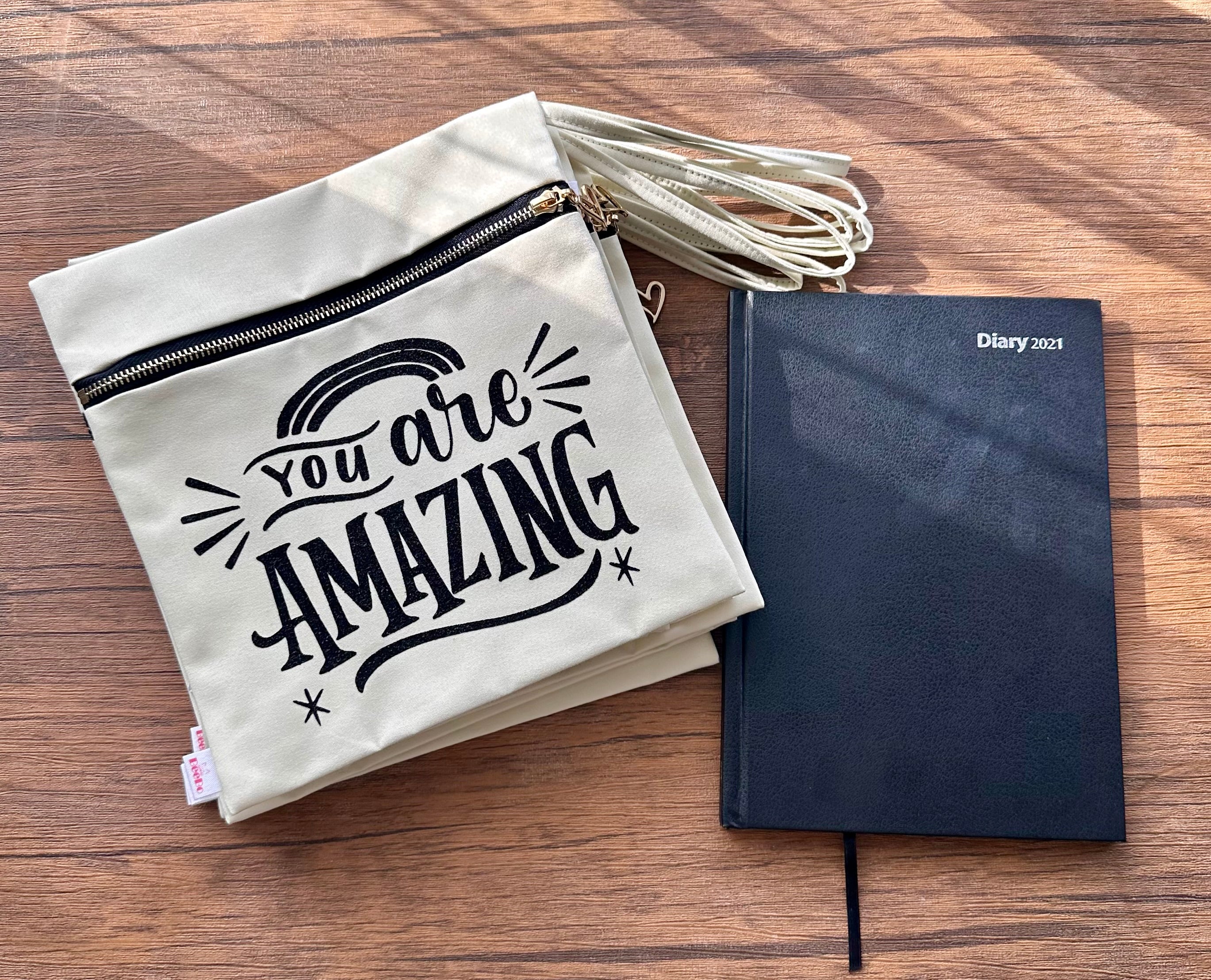 Beige pouch with 'You are Amazing' text and black notebook on a wooden surface