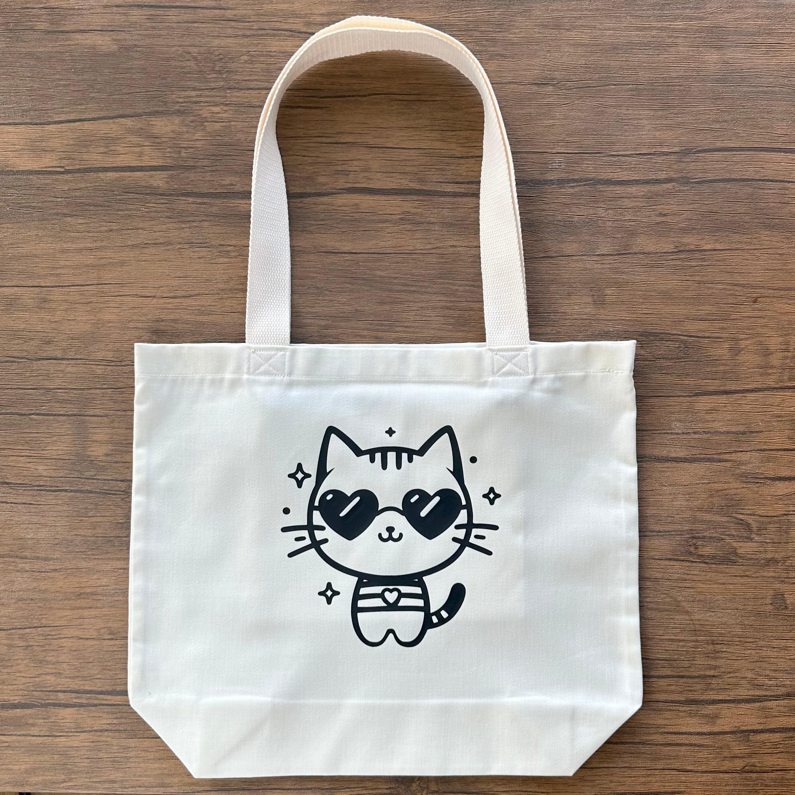 White tote bag with a black cat illustration wearing sunglasses on a wooden surface