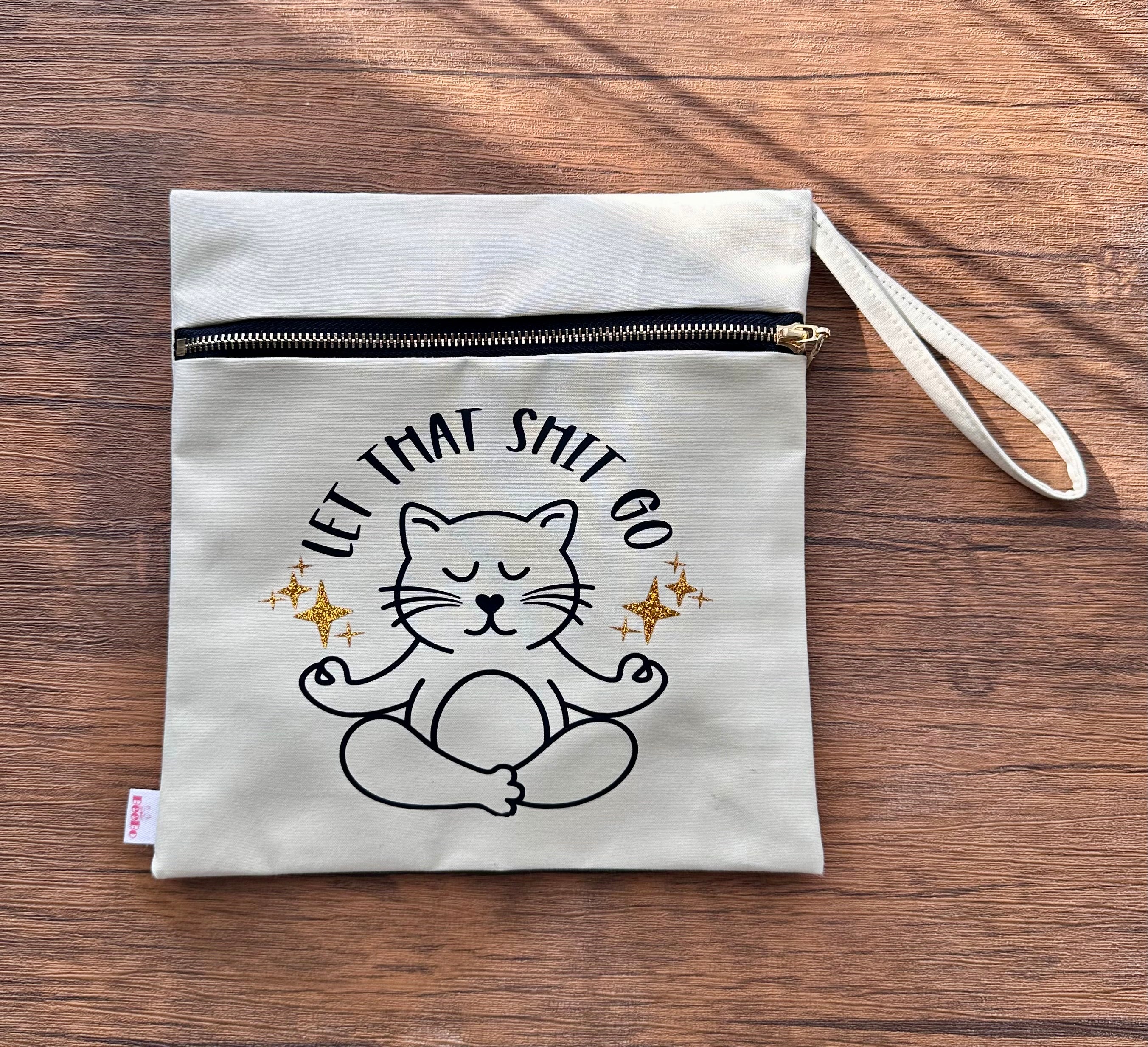 Canvas pouch with a cat illustration and text 'Let That Shit Go' on a wooden surface