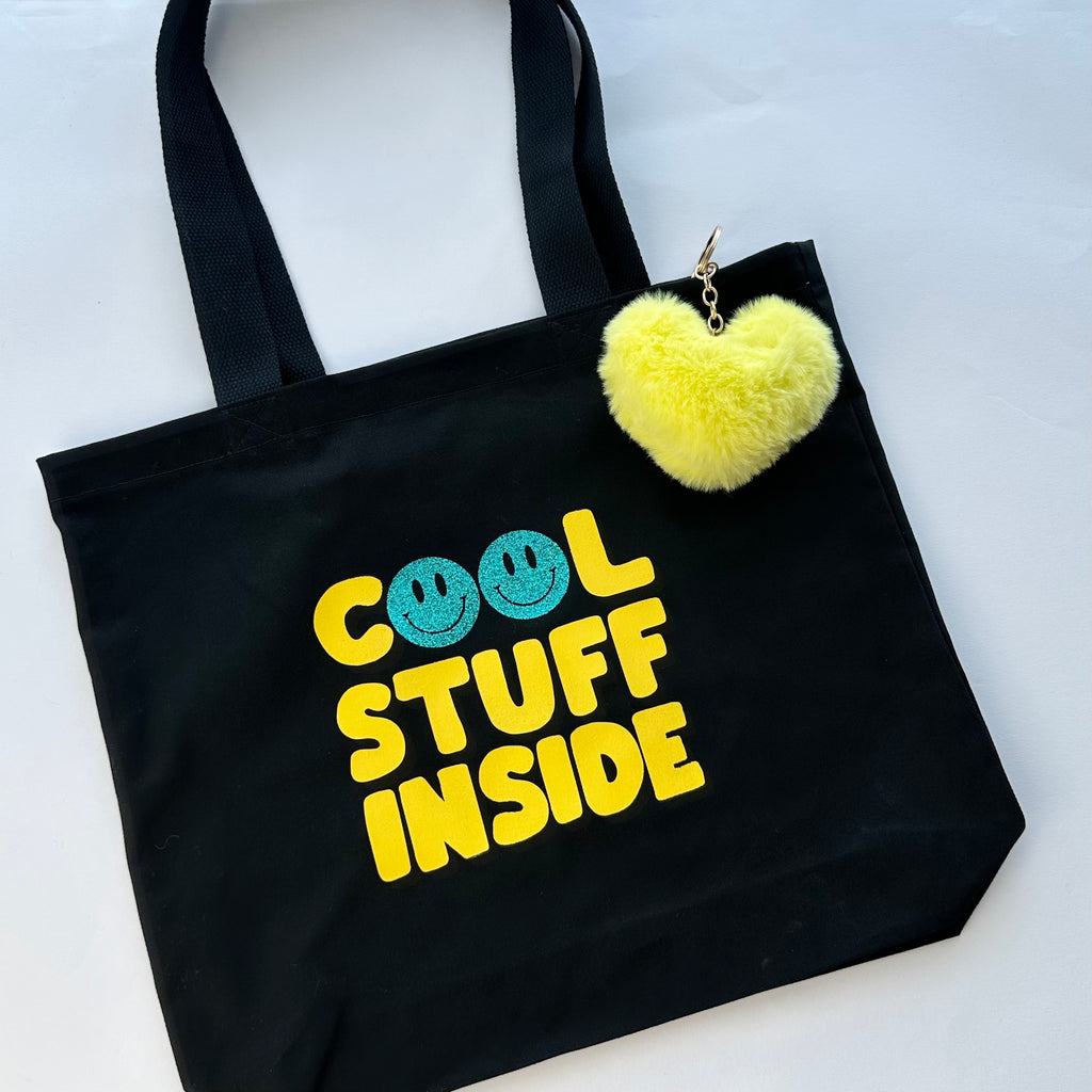 Black tote bag with 'Cool Stuff Inside' text and a yellow heart-shaped pom-pom keychain on a white background