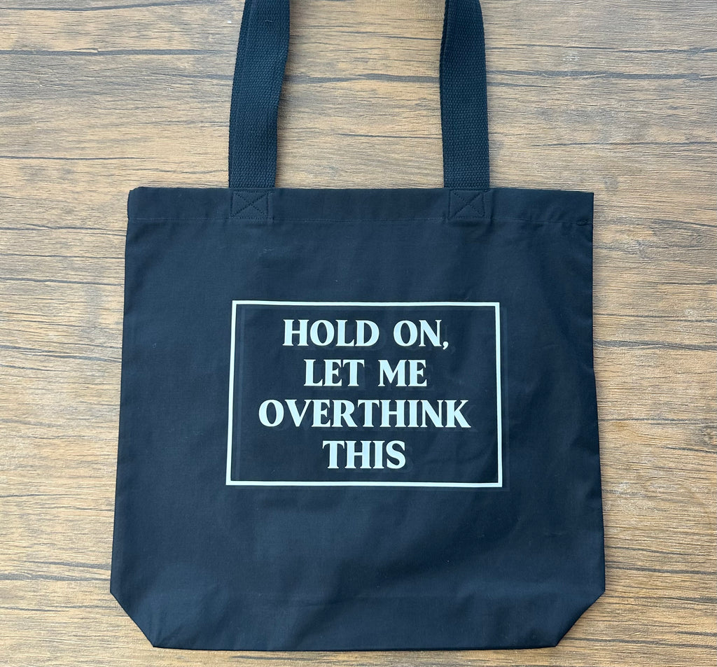 Black tote bag with text 'Hold on, let me overthink this' on a wooden surface
