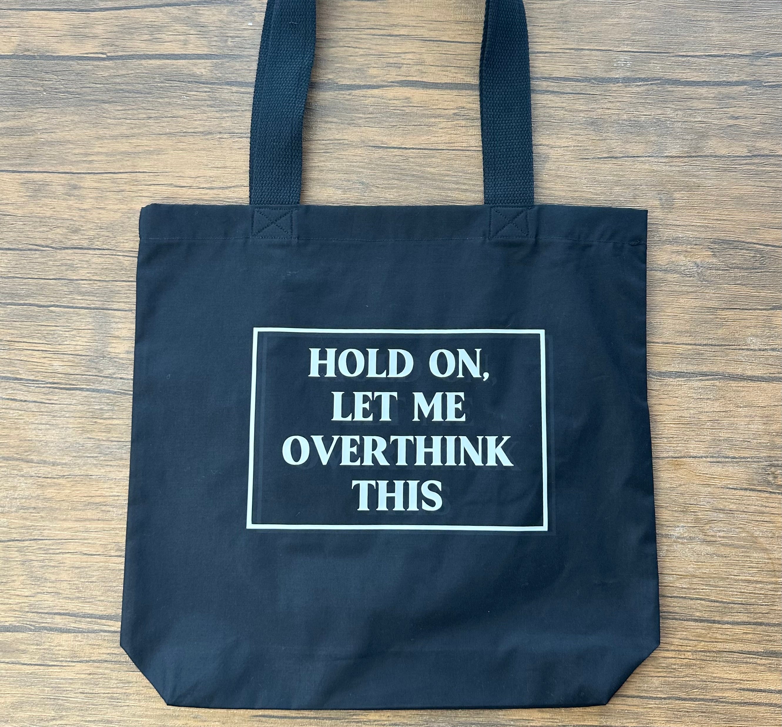Black tote bag with text 'Hold on, let me overthink this' on a wooden surface