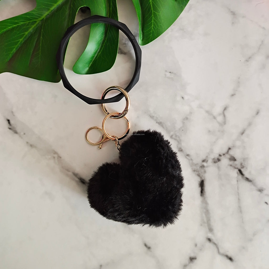Black pom-pom keychain with gold rings on a marble surface with green leaves.
