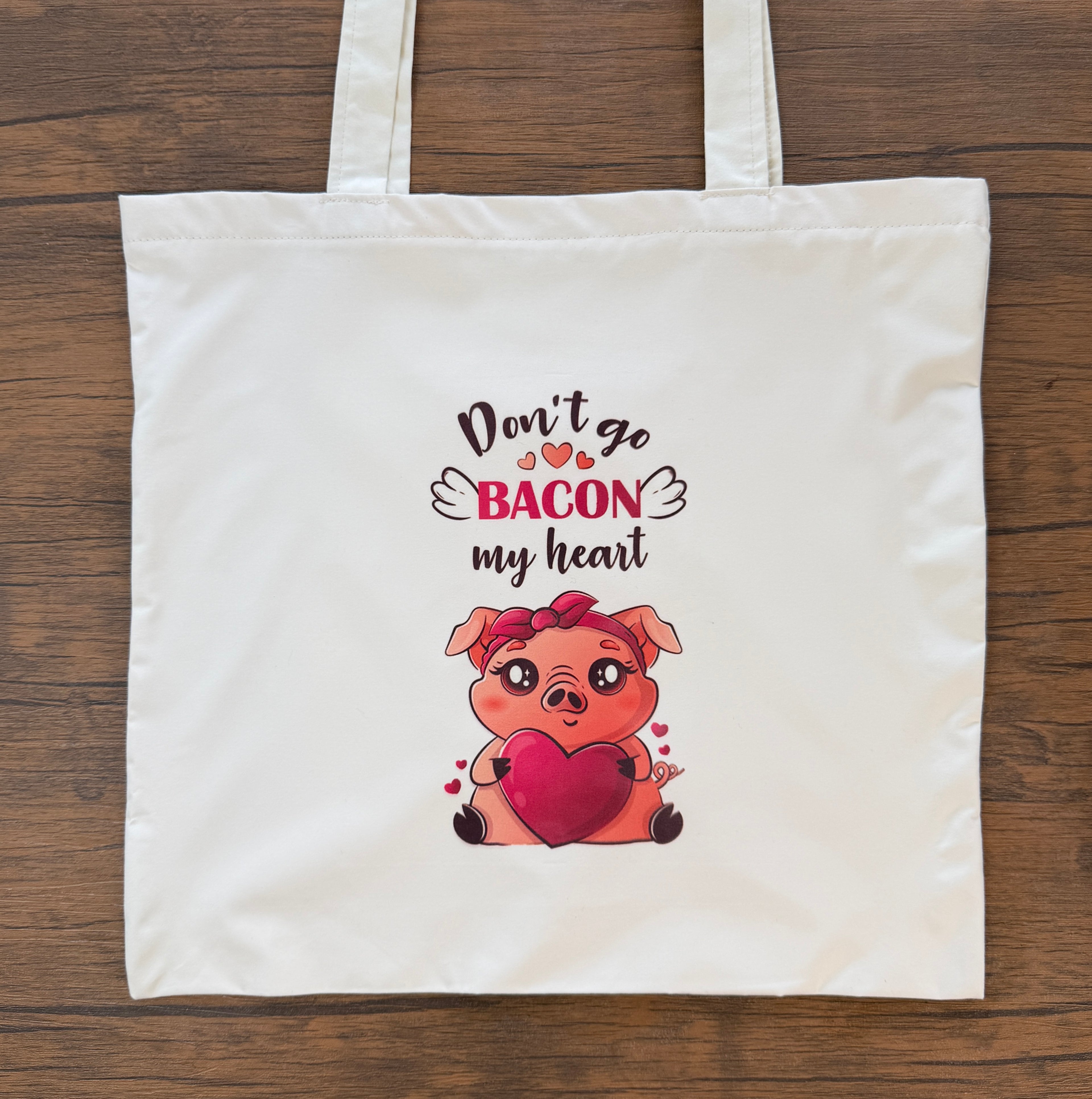 Tote bag with a cartoon pig holding a heart and text 'Don't go bacon my heart' on a wooden surface.