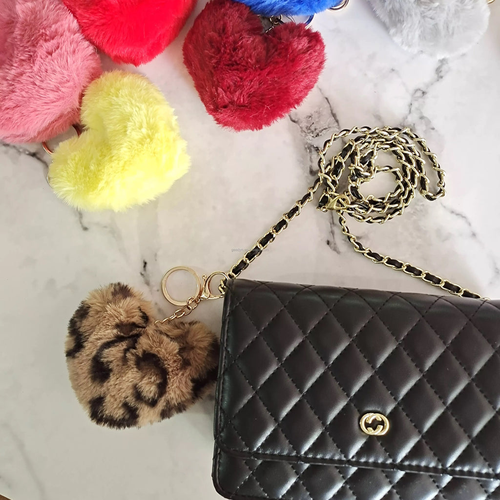 Black quilted handbag with gold chain strap and colorful pom-pom keychains on a marble surface